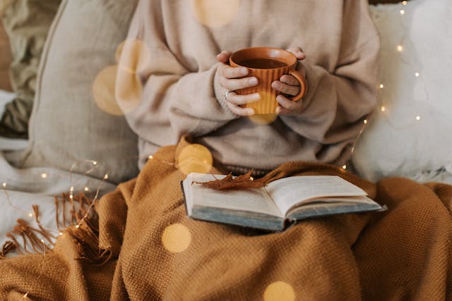 A person covered with a blanket reading a book and drinking tea.